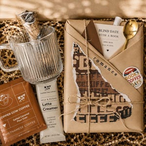May include: A gift box containing a ribbed glass mug, a packet of Copper Cow Coffee salted caramel pour over, a Copper Cow Coffee latte creamer packet, and a book wrapped in brown paper with a tag reading Blind Date With A Book.
