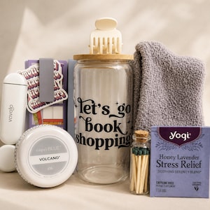 May include: A gift set featuring a clear glass tumbler with the text let's go book shopping, a white hair claw clip, a gray fuzzy towel, a Capri Blue Volcano body butter, a small bottle of matches, a box of Yogi Honey Lavender tea, and a white device.