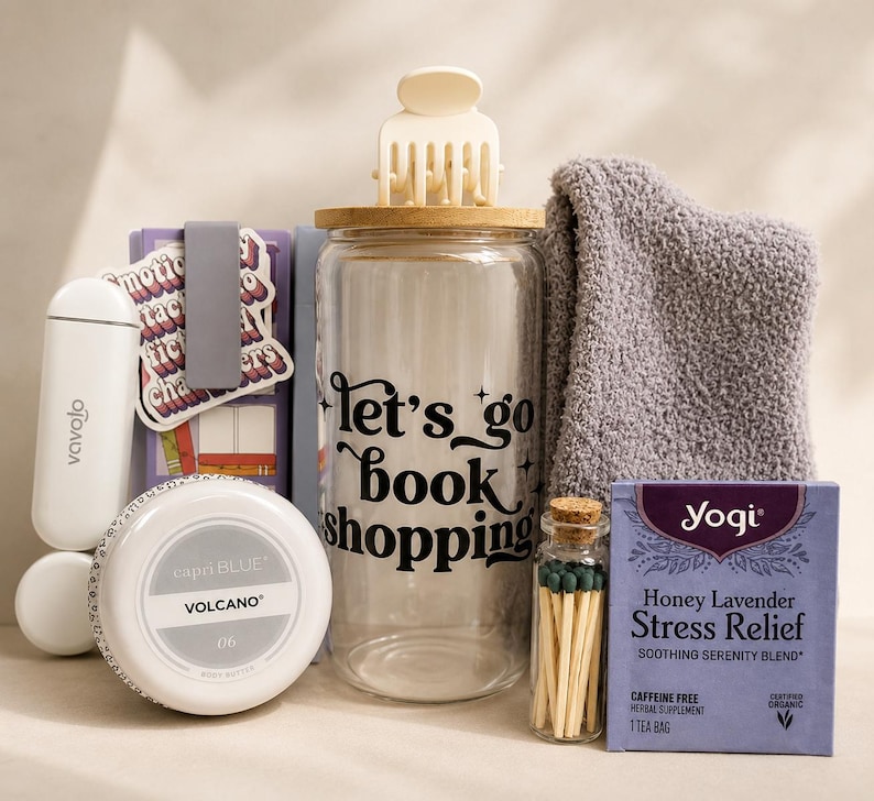 May include: A gift set featuring a clear glass tumbler with the text let's go book shopping, a white hair claw clip, a gray fuzzy towel, a Capri Blue Volcano body butter, a small bottle of matches, a box of Yogi Honey Lavender tea, and a white device.