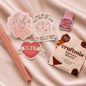 May include: A peach-colored marker, three die-cut stickers, a small pink magnetic bookmark, and a Craftmix Espresso Martini drink packet. The stickers feature text including Light it up, Roses are red these pages are worn this book I'm reading is basically porn, and Smut. The bookmark reads Smutty Book Club.