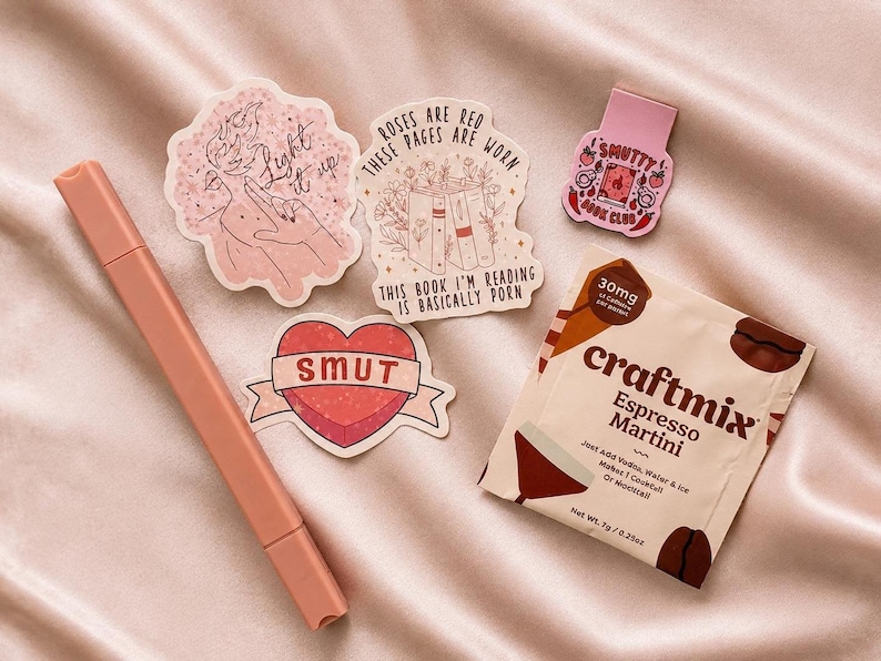 May include: A peach-colored marker, three die-cut stickers, a small pink magnetic bookmark, and a Craftmix Espresso Martini drink packet. The stickers feature text including Light it up, Roses are red these pages are worn this book I'm reading is basically porn, and Smut. The bookmark reads Smutty Book Club.