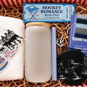 May include: A gift box containing a white towel with hockey stickers, a frosted glass tumbler with a bamboo lid, a light blue marker, a blue Hockey Romance Book Club ticket, a Comfort soak packet, and a black circular hockey-themed coaster.