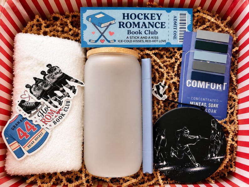 May include: A gift box containing a white towel with hockey stickers, a frosted glass tumbler with a bamboo lid, a light blue marker, a blue Hockey Romance Book Club ticket, a Comfort soak packet, and a black circular hockey-themed coaster.