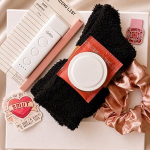 May include: A reading list card, a white Gritin book light, a pink pen, black fuzzy socks, a red tea packet, a pink satin scrunchie, and two stickers. One sticker is a heart with the text SMUT, and the other is a pink rectangle with SMUTTY BOOK CLUB.