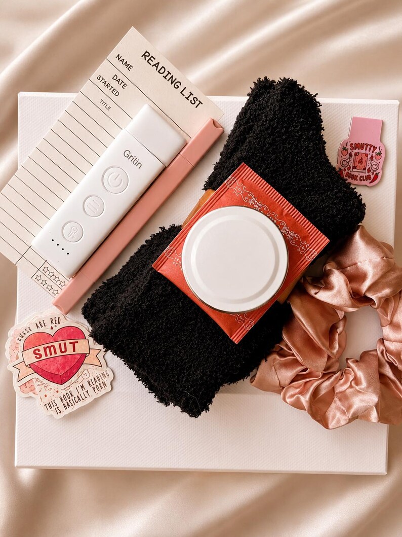 May include: A reading list card, a white Gritin book light, a pink pen, black fuzzy socks, a red tea packet, a pink satin scrunchie, and two stickers. One sticker is a heart with the text SMUT, and the other is a pink rectangle with SMUTTY BOOK CLUB.