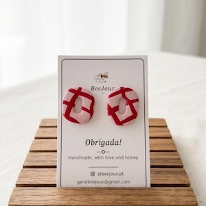 Op de afbeelding: Een paar ovale oorbellen met een lichtroze basis en een opvallend rood ruitpatroon. De oorbellen worden gepresenteerd op een witte kaart met het label BeeJoux en de tekst Obrigada! Handmade, with love and honey, @beejoux.pt en geralbeejoux@gmail.com.