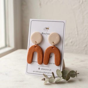 Peut inclure: Boucles d'oreilles pendantes composées d'une pièce supérieure beige circulaire reliée à une pièce inférieure plus grande en forme d'arche de couleur terre cuite. Présentées sur une carte blanche avec le texte BeeJou et des coordonnées.