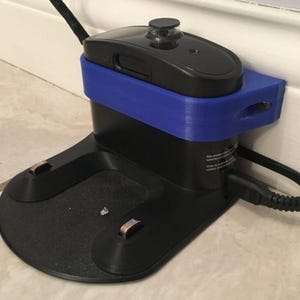 May include: A black robotic vacuum charging dock featuring a custom-fitted blue plastic bracket attached to the back. The dock has a flat base with two metal charging contacts and a power cord plugged into the side, positioned against a white wall.