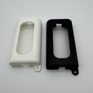 May include: Two rectangular protective covers, one in white and one in black, featuring a central elongated cutout and a small circular mounting tab at the bottom. These 3D printed plastic housings have a textured surface and rounded corners.