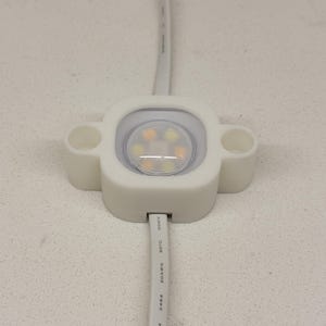 May include: A white square LED module with rounded corners and two side mounting holes, featuring a central circular lens containing multiple small light points. A white electrical wire extends from the top and bottom of the housing.