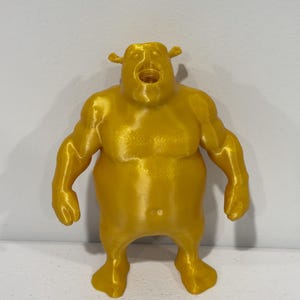 Op de afbeelding: Een 3D-geprint goudgeel beeldje van een stevig, gespierd mensachtig figuur met grote oren en een wijd open mond. Het figuur heeft een glad, reflecterend oppervlak en staat rechtop met de armen langs het lichaam uitgestrekt.
