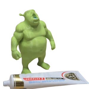 Op de afbeelding: Een lichtgroen, 3D-geprint ogre-figuurtje met een wijde, open mond, staand achter een witte tube Complex 7 Total Care Plus tandpasta. Het figuurtje heeft een stevig, gespierd postuur en opvallende oren, geplaatst naast het tandverzorgingsproduct.