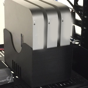 May include: A black plastic vertical storage stand holding three silver square computer units in a row. The stand features a curved cutout on the side and vertical ventilation slots. Each silver computer unit has a small circular status light on the front.