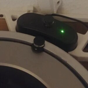 May include: A circular silver metallic device with a black knob on its edge, positioned next to a black rectangular control unit featuring a single green LED light. The control unit is mounted on a white triangular bracket with a geometric cutout design.