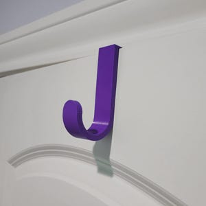 May include: A purple over-the-door hook with a flat vertical back and a curved J-shaped front. The hook is constructed from a solid material and is positioned securely over the top edge of a white paneled door, extending downward to provide storage.
