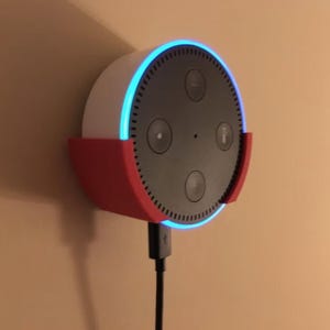 May include: A circular smart speaker with a black front panel and white casing, mounted in a red plastic wall bracket. The device features a glowing blue light ring around the perimeter and is connected to a black power cable at the bottom.