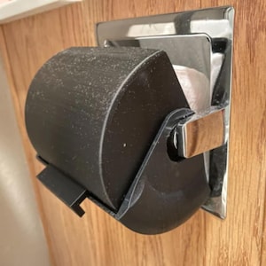 May include: A black, curved, 3D-printed toilet paper roll cover mounted onto a rectangular, polished chrome metal wall fixture. The cover features a smooth, matte finish and a small protruding tab at the bottom edge, partially obscuring a white paper roll.