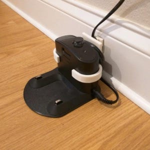 May include: A black robotic vacuum charging dock station featuring a white 3D-printed wall mount bracket. The dock has a circular base with two metal charging contacts and a black power cord connected to the side, plugged into a wall outlet.