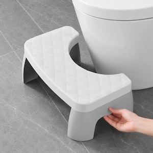 May include: A light grey plastic toilet stool featuring a curved cutout design and a white top surface with a quilted diamond pattern. The stool has a sturdy base and is designed to fit around the base of a standard toilet for foot support.