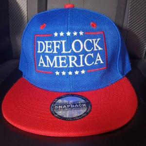 May include: A blue snapback cap with a red brim and a red button on top. The front features a rectangular red border enclosing the white embroidered text DEFLOCK AMERICA, flanked by five white stars above and five white stars below the text.