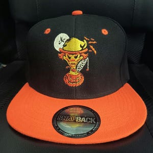 May include: A black snapback cap with an orange brim and an orange button on top. The front features an embroidered graphic of a yellow jester head emerging from an orange jack-o'-lantern, set against a white moon. A circular sticker reads SNAPBACK.
