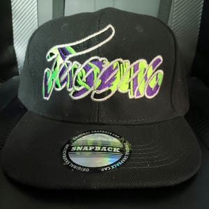 May include: A black snapback cap featuring an embroidered logo on the front with stylized lettering in shades of purple and neon green. A circular holographic sticker is affixed to the flat brim, displaying the text Original Snapback Cap.