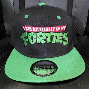 May include: A black snapback cap with a bright green brim and a green button on top. The front features embroidered text that reads I AM ACTUALLY IN MY FORTIES in a stylized font with a red rectangular background behind the first line of text.