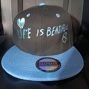 May include: A black snapback cap with a light blue brim and a light blue button on top. The front features embroidered light blue text that reads LIFE IS BEAUTIFUL, a broken heart icon, and a circular symbol. A round holographic sticker is on the brim.