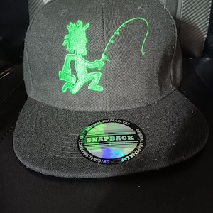 Könnte beinhalten: Eine schwarze Snapback-Cap mit einer leuchtend grünen Stickerei, die eine Figur mit einer Angelrute zeigt. Ein runder Aufkleber auf dem flachen Schirm zeigt den Text ORIGINAL SNAPBACK CAP und SNAPBACK in weißer und grüner Schrift.