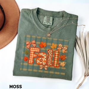 May include: A moss green short-sleeve t-shirt with a crew neckline. The front features a graphic with the word Fall in orange and white patterns, surrounded by orange maple leaves, bows, and pumpkins on a green plaid background.