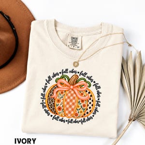 May include: An ivory t-shirt featuring a central graphic of a pumpkin with a peach-colored bow. The pumpkin has sections with leopard print, orange gingham, and floral patterns. The text "fall vibes" repeats in a circle around the pumpkin.