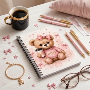 Può includere: Un quaderno a spirale con un orsetto marrone che indossa un maglione e un fiocco rosa, che tiene una tazza con la scritta LOVELY. La copertina presenta stelle e fiocchi rosa. Testo: COLLEZIONE COZY TEDDY, RENDI OGNI GIORNO PIÙ DOLCE, IL TUO QUADERNO, LA TUA ISPIRAZIONE.