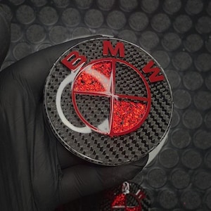 Puede incluir: Un emblema circular de BMW con un fondo de patrón de fibra de carbono negro. El logo incluye las letras BMW en rojo y un diseño circular central dividido en cuatro cuadrantes, con dos secciones rellenas de purpurina roja vibrante.