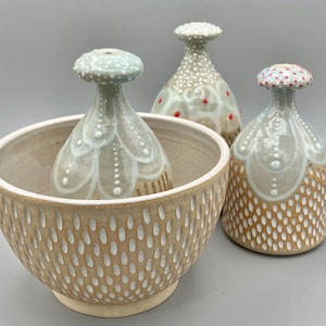 May include: A collection of handmade ceramic pieces. The set includes a large beige bowl with a dotted pattern, and three decorative bottles with mushroom-shaped tops. The bottles have a light green and white color scheme with dotted and floral designs.