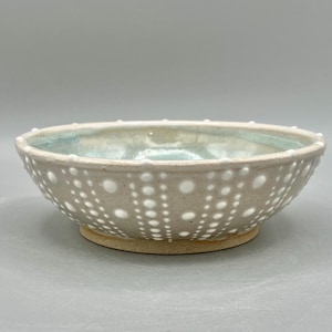 May include: A handmade ceramic bowl with a light beige exterior and a pale blue interior. The bowl features a pattern of evenly spaced white dots. The bowl is approximately 6 inches in diameter and 3 inches tall.