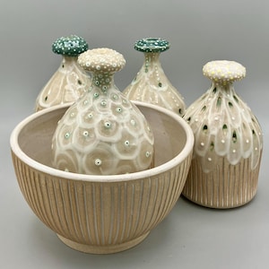 May include: A collection of handmade ceramic pieces. The image features a beige bowl with vertical lines and four decorative mushroom-shaped vases. The vases have a beige base with white and green accents, and a dotted pattern.