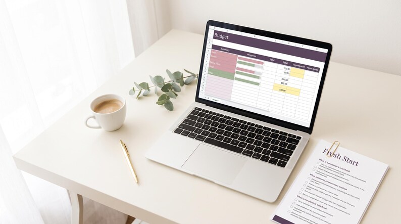 May include: A silver laptop displaying a budget spreadsheet with purple and green accents, a white coffee mug, a gold pen, and a document titled Fresh Start with a gold paperclip, all arranged on a clean, light-colored surface.