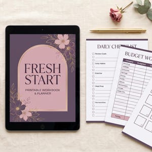 May include: A tablet displaying a digital cover titled Fresh Start Printable Workbook & Planner with floral graphics, alongside printed sheets for a daily checklist, budget worksheet, and savings tracker, a gold pen, and a pink mug.