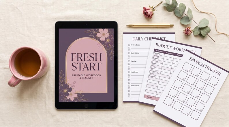 May include: A tablet displaying a digital cover titled Fresh Start Printable Workbook & Planner with floral graphics, alongside printed sheets for a daily checklist, budget worksheet, and savings tracker, a gold pen, and a pink mug.