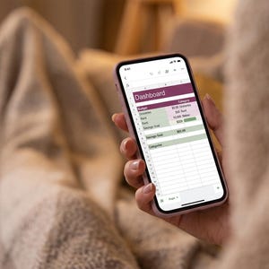 May include: A smartphone displaying a digital budget dashboard on its screen. The interface features a header labeled Dashboard, with rows for categories like Groceries, Rent, and Savings Goal, alongside numerical data and progress bars in shades of green.