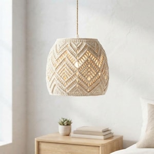 Op de afbeelding: Een crèmekleurige hanglamp met een cilindrische kap gemaakt van ingewikkeld macramékoord. De kap heeft een herhalend geometrisch chevronpatroon en hangt aan een enkel dun koord dat aan het plafond is bevestigd.