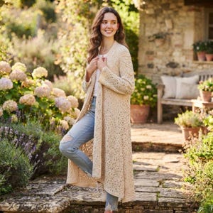 May include: A long, cream-coloured open-front cardigan featuring an intricate, open-knit lace pattern with long sleeves, paired with light blue distressed denim jeans that have a visible rip at the knee and a simple white undershirt.