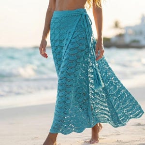 May include: A floor-length turquoise crochet wrap skirt featuring a scalloped pattern throughout the fabric. The skirt includes a matching crocheted tie closure at the waist with long, dangling tassels that hang down the side of the garment.