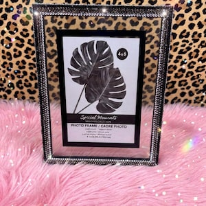 May include: A rectangular photo frame with a border encrusted in small, shimmering silver rhinestones. The frame holds a display card featuring a black and white illustration of two monstera leaves and the text 4x6, Special Moments, and Photo Frame.