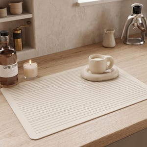 May include: A rectangular beige silicone drying mat with horizontal grooves, featuring a matching beige ceramic mug and a rounded saucer. The mat is placed on a wooden surface next to a glass bottle of coffee syrup and a lit candle in a glass jar.