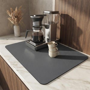 Peut inclure: Un tapis de cuisine rectangulaire gris foncé aux coins arrondis posé sur un comptoir en marbre. Une cafetière argentée avec une carafe en verre remplie de café noir est posée sur le tapis, accompagnée d'une pile de trois petites tasses en céramique beige.
