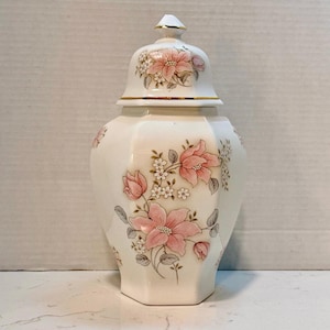 Pode incluir: Um pote de gengibre de porcelana branca com tampa removível, apresentando um corpo hexagonal e um padrão floral de flores cor-de-rosa, folhas verdes e pequenas flores brancas. A tampa é rematada com um detalhe dourado.