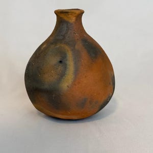 Puede incluir: Un pequeño jarrón de arcilla de forma redonda, moldeado a mano, con un cuello estrecho y un cuerpo ancho y bulboso. La superficie presenta una paleta de colores terrosos y moteados de naranja quemado, marrón oscuro y gris carbón.