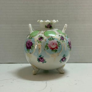 May include: A small, round, white porcelain vase with three feet and two handles. The surface features hand-painted pink and purple roses set within light blue circular medallions, accented by green scalloped details and delicate gold filigree.