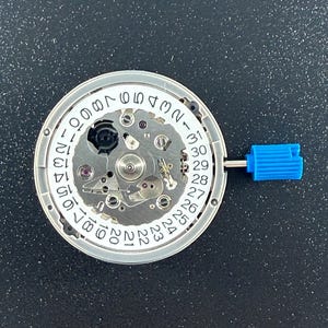 May include: A circular mechanical watch movement featuring a white date wheel with black numerals from 1 to 31 printed around the perimeter. The central mechanism displays metallic gears, screws, and a black component, with a blue plastic crown stem.
